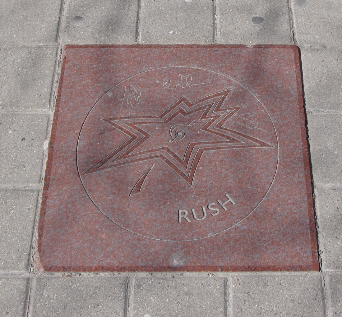 A red granite star set into a sidewalk, carved with a star, a treble clef, the word RUSH and three signatures.